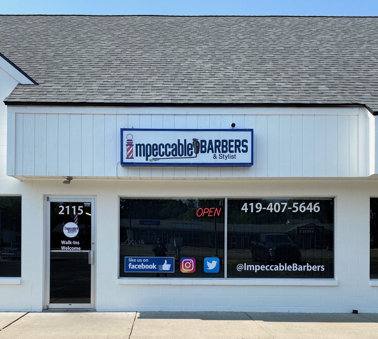 The front of Impeccable Barbers and Stylist: a white storefront on North Reynolds Road with a large blue and white sign reading impeccable BARBERS and Stylist, a red neon OPEN sign in the window, the phone number 419-407-5646, and a glass door marked 2115 with Walk-Ins Welcome.