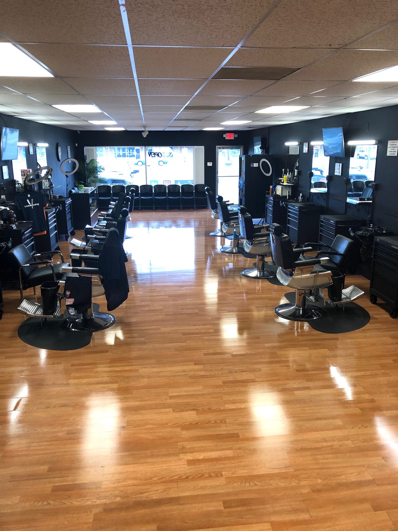 Inside the shop looking toward the front window: barber chairs on both sides, a row of waiting chairs by the front window, daylight coming through the glass, and stations with mirrors and tool drawers.